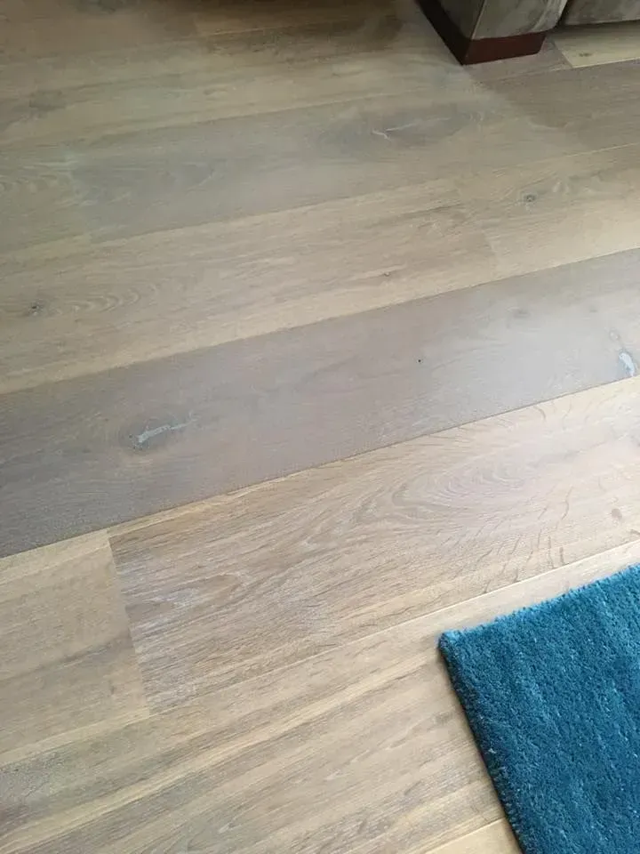 A close-up view of light-toned engineered wood flooring featuring planks with varying grain patterns and a blue rug corner.