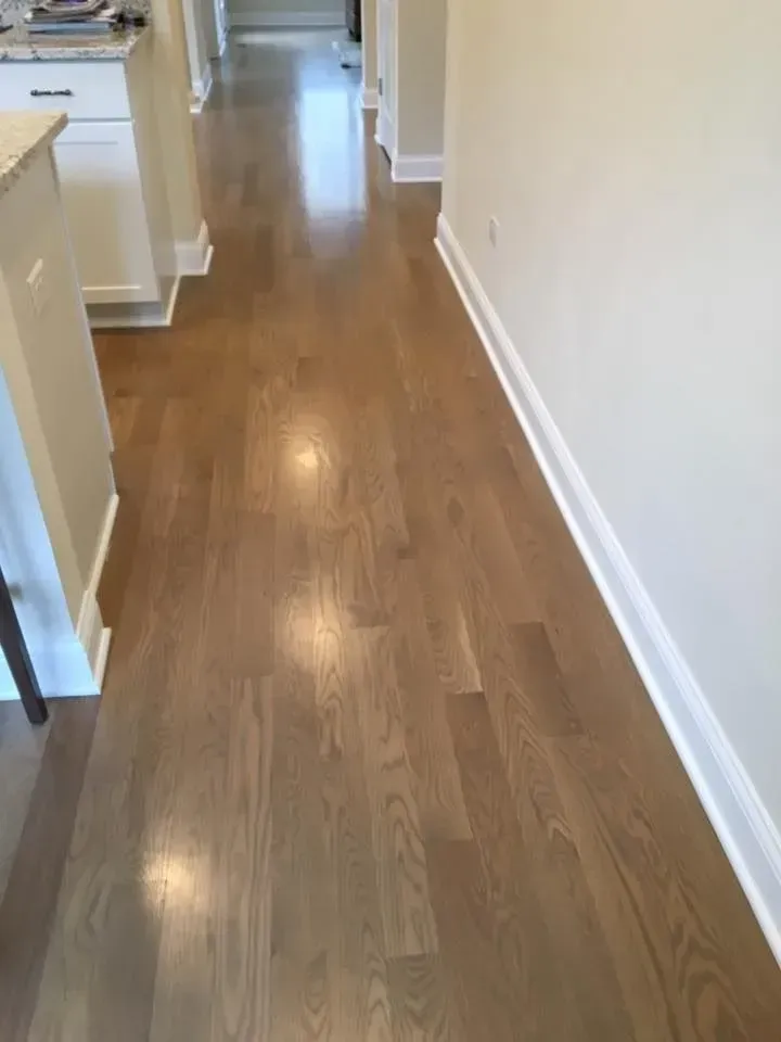 A hallway with light-brown hardwood flooring, white baseboards, and neutral-colored walls.