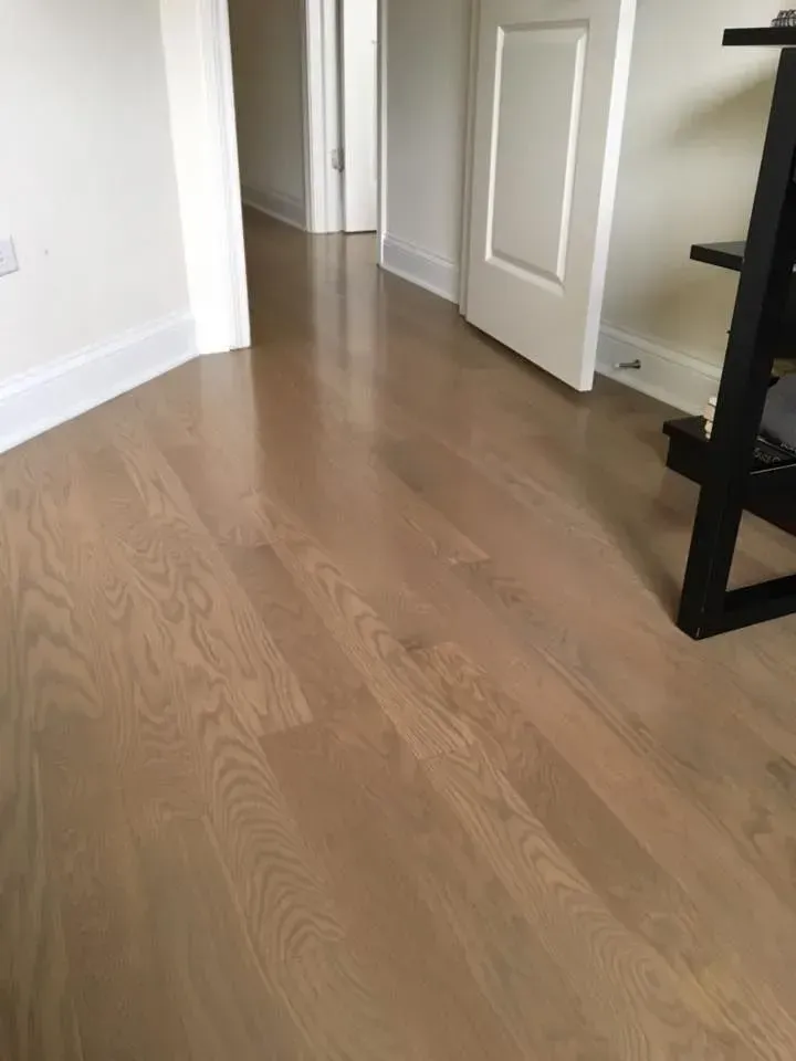 Light-toned, polished hardwood flooring in a room with white trim, a hallway, and a partially visible black shelf.
