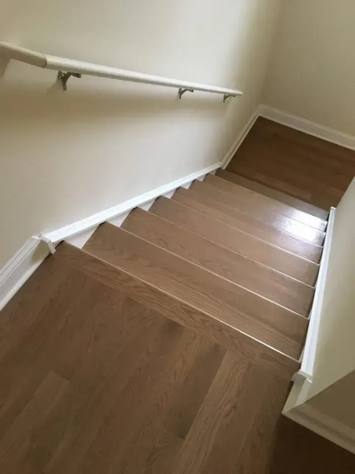 A downward view of a wooden staircase with a white wall, white trim, and a white handrail.