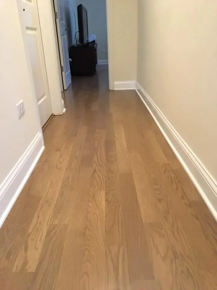 A hallway with light oak hardwood flooring, white baseboards, and beige walls leading into an adjacent room.