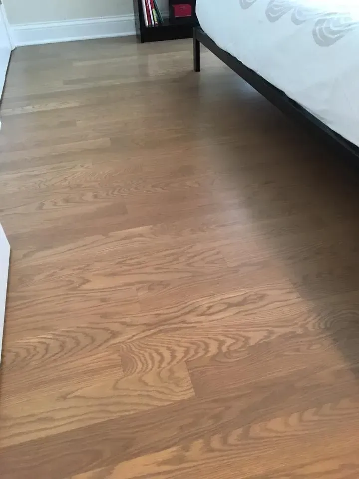 A close-up view of light-brown hardwood flooring in a room, with the base of a bed frame visible in the background.