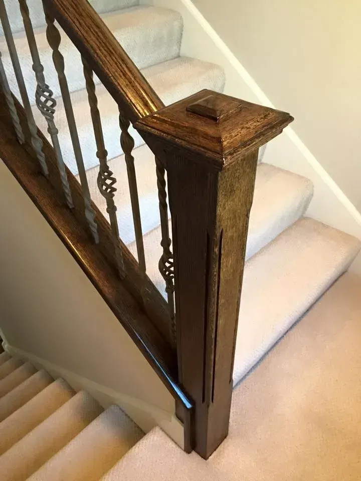 Wooden staircase with a dark handrail, decorative metal balusters, and a newel post with a faded finish on carpeted stairs.