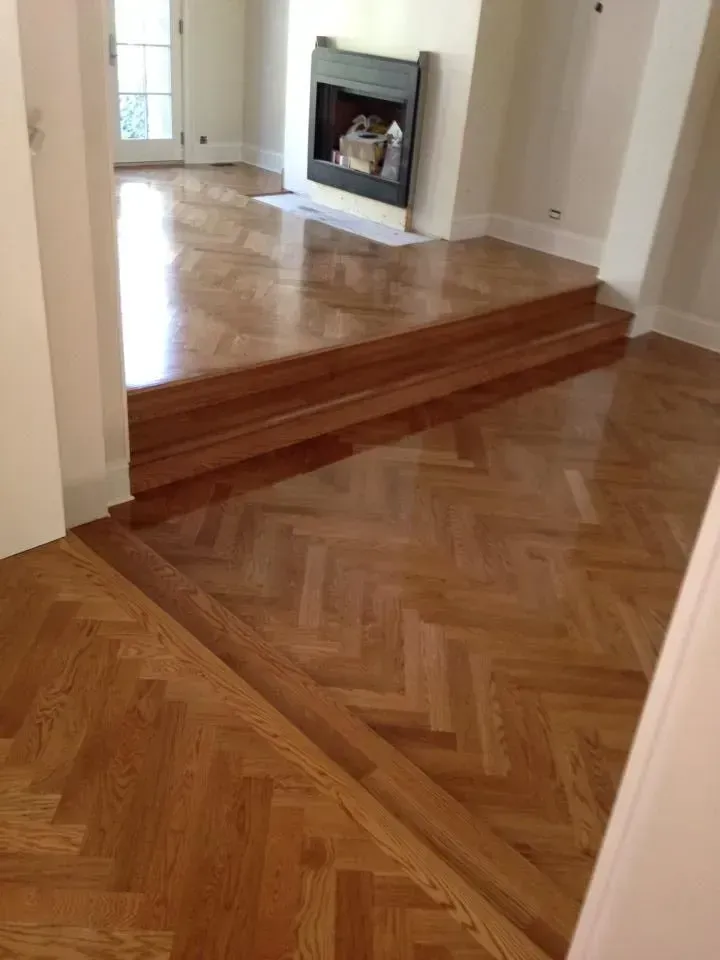 Herringbone hardwood floors in a light brown finish transition between a sunken living area and a raised fireplace section.
