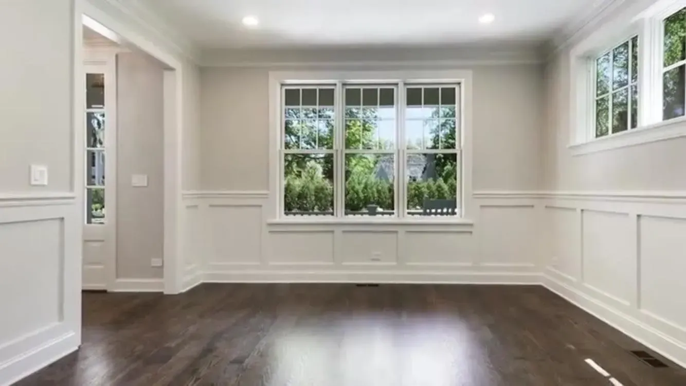 A bright dining room with white wainscoting, light gray walls, dark hardwood floors, and a large central window.