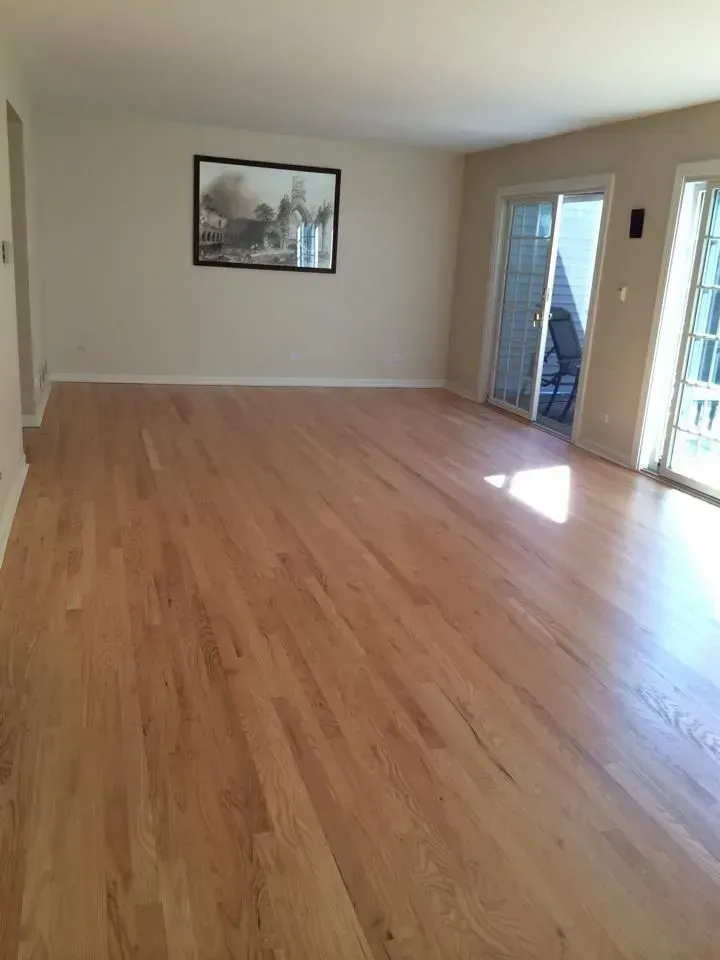 An empty room with light-colored hardwood floors, beige walls, a framed black-and-white picture, and glass sliding doors.