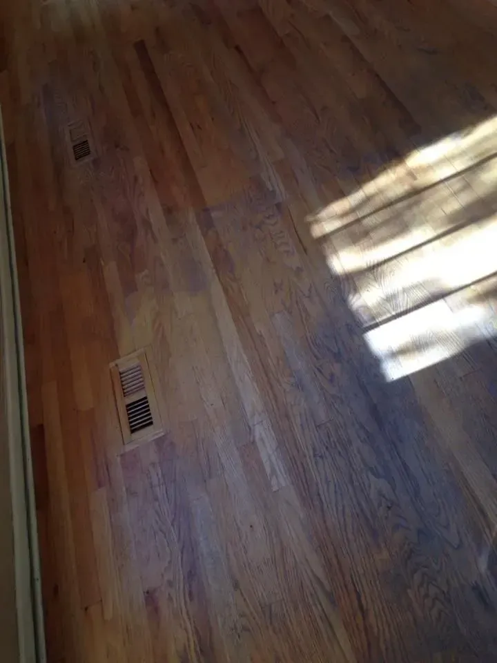 Close-up of a hardwood floor with a metal ventilation grate and bright sunlight reflecting on the surface.