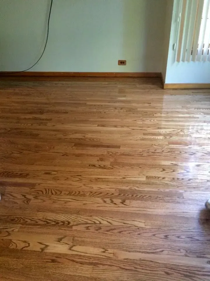 A bright, empty room featuring polished medium-brown wood flooring, light-colored walls, and vertical window blinds.