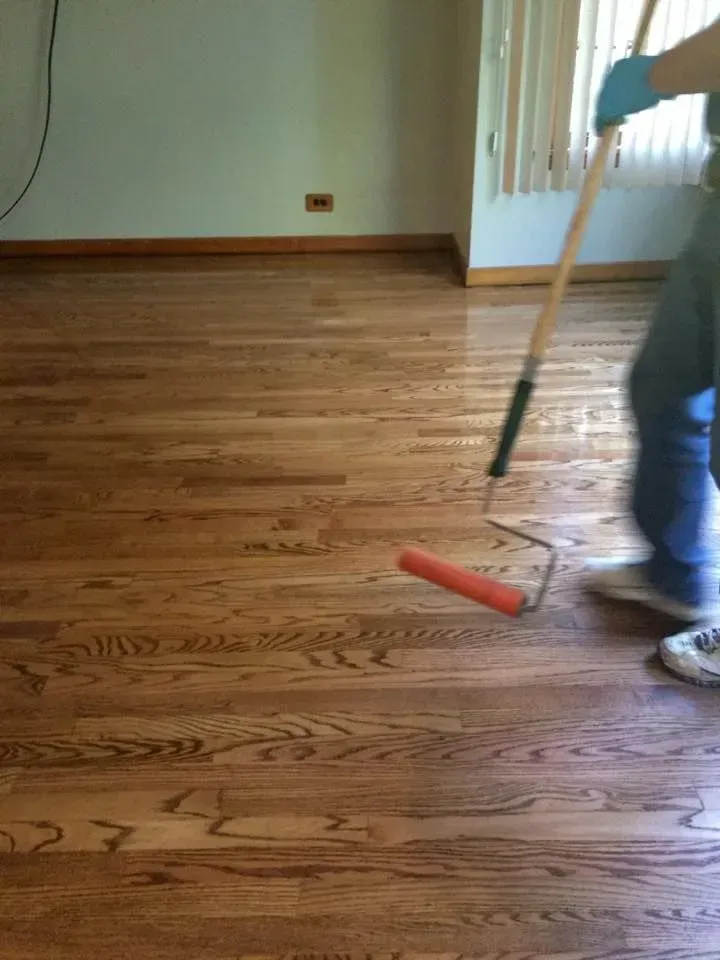 A person in blue gloves uses a long-handled paint roller to apply a clear finish to a hardwood floor in a room.
