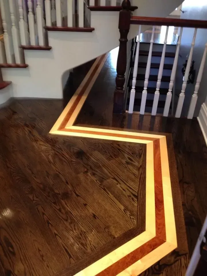 A zigzag border of light and reddish wood accents a dark wood floor near a staircase with white railings.
