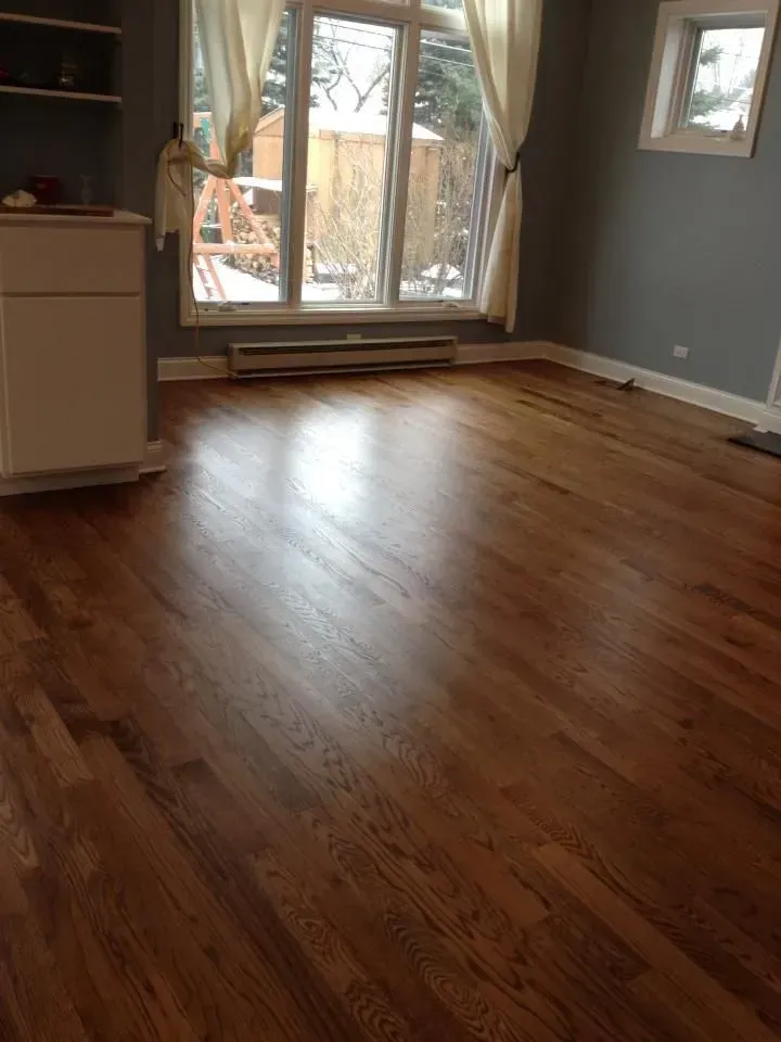 An empty room with polished hardwood floors, grey walls, white cabinets, and large windows with sheer curtains.