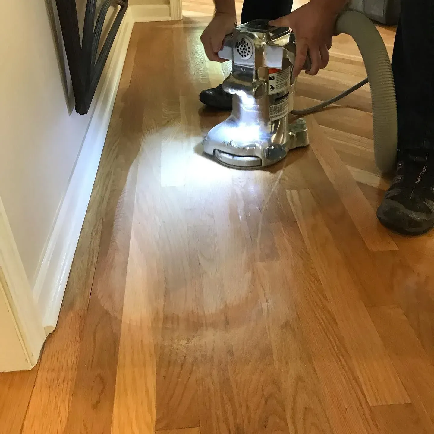 A person uses a floor sander with a vacuum attachment to refinish wood flooring near a white baseboard.