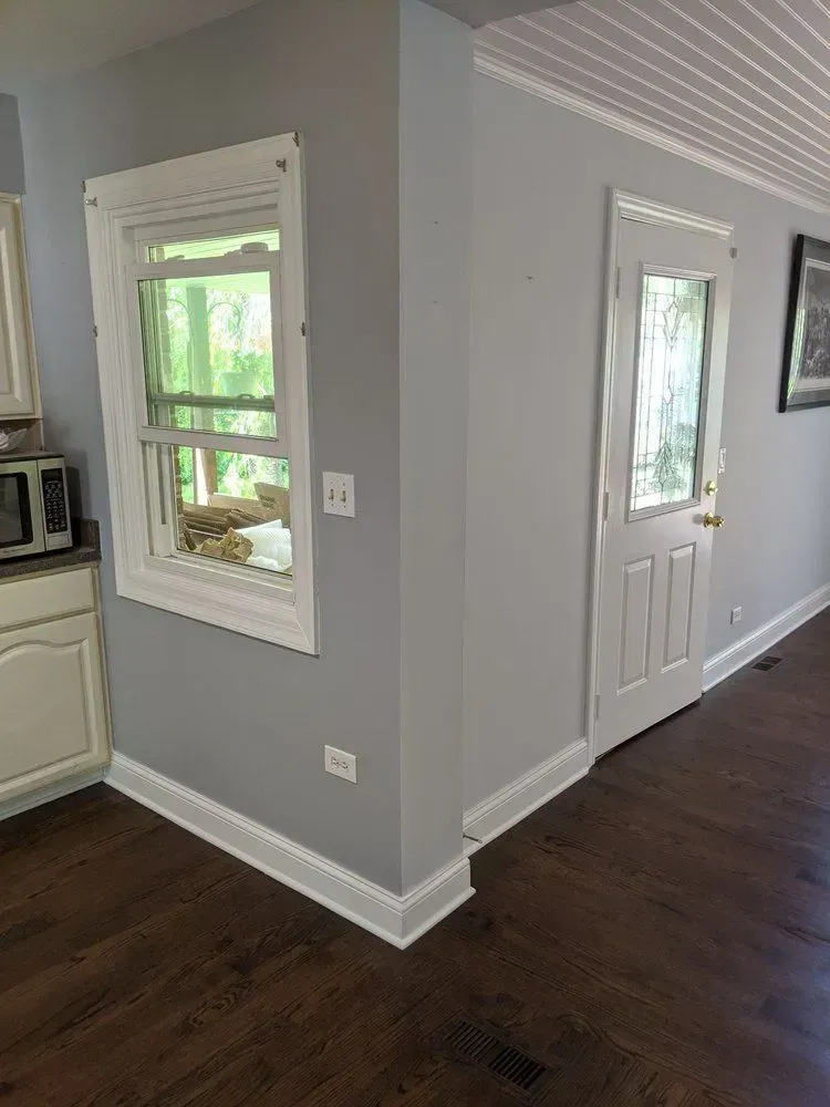A corner of a room featuring a window with white trim, a white door, light gray walls, and dark wood flooring.