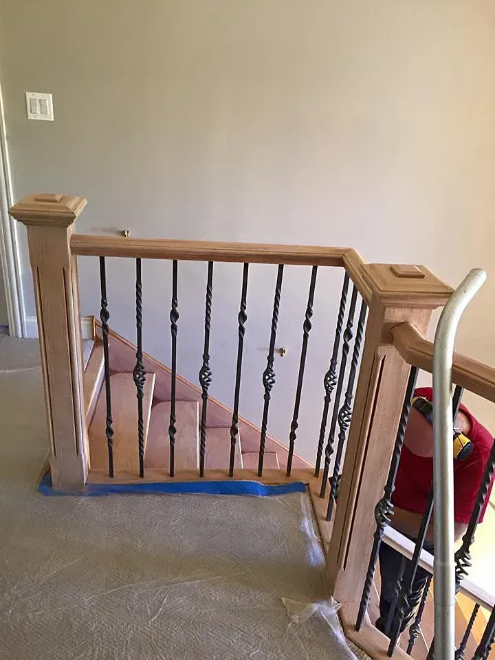 A wooden staircase railing with black iron balusters under construction, with blue painter's tape on the floor.