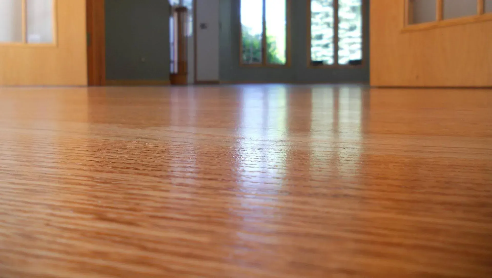 Close-up of polished light brown wood flooring in a room with a blurred background showing doors and windows.