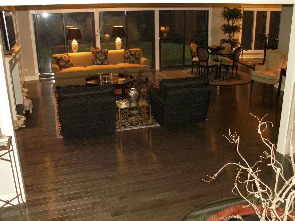 A living room with dark hardwood floors, two black armchairs, a tan sofa, and a dining set near large sliding glass doors.