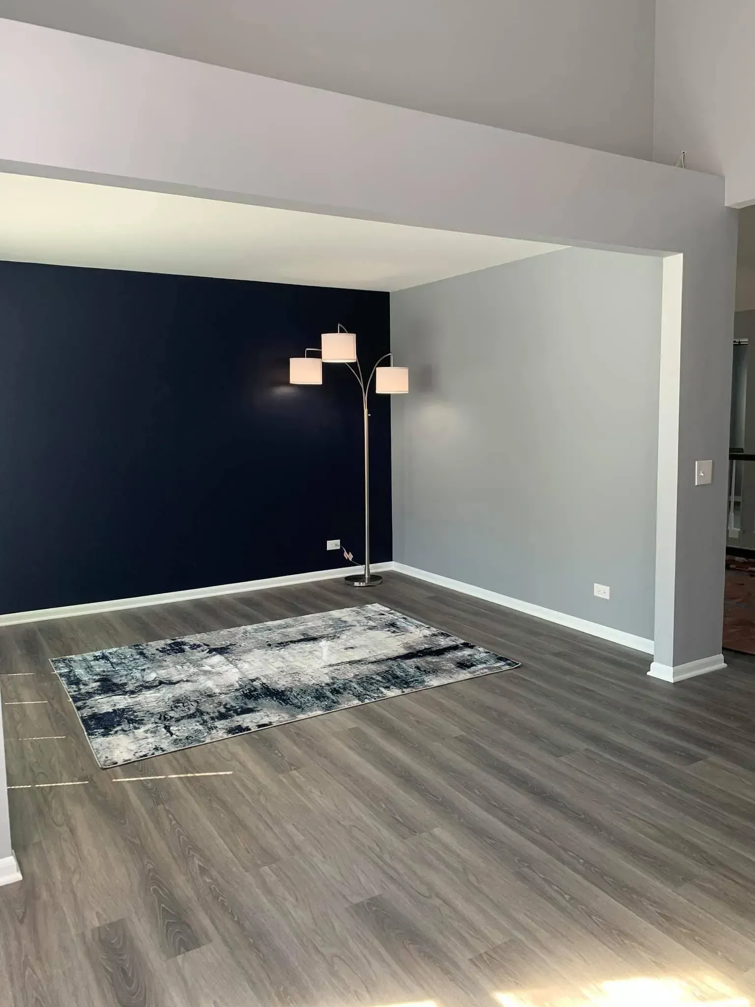 A modern living area with dark blue and gray walls, wood-look flooring, a patterned area rug, and a three-bulb floor lamp.