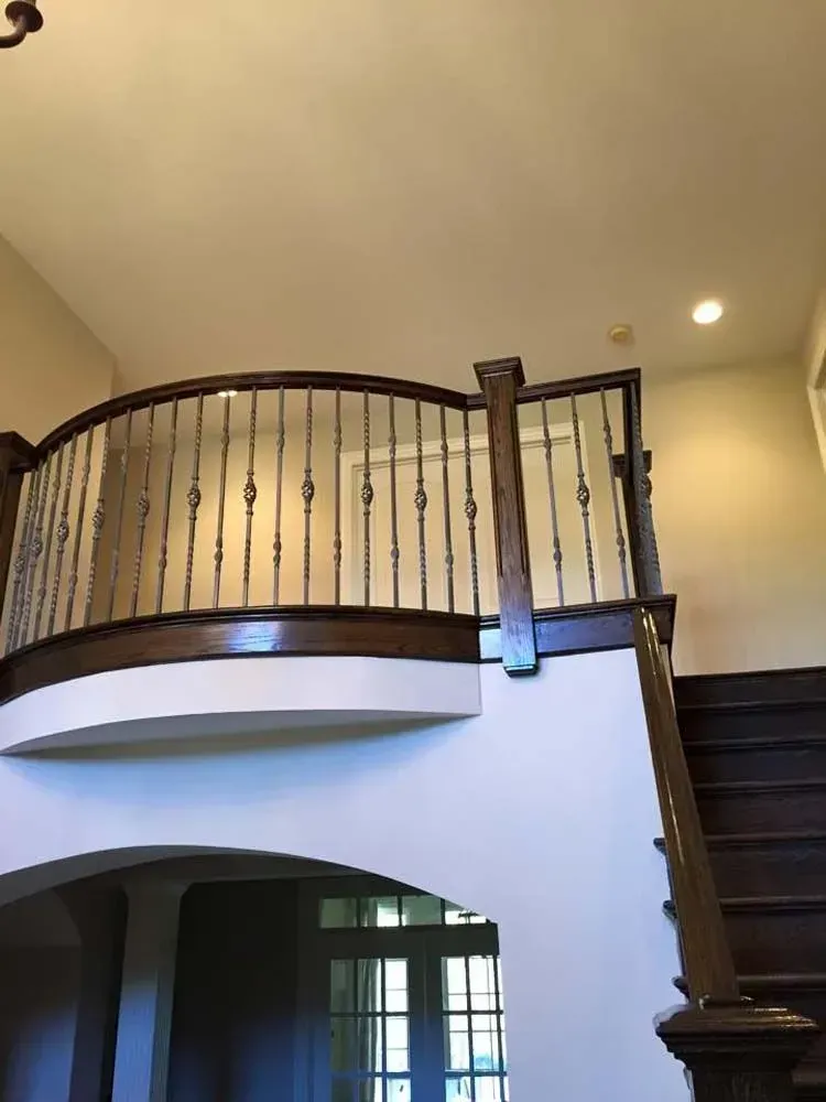 A curved wooden balcony railing with metal balusters overlooking an entryway with stairs and an arched doorway.