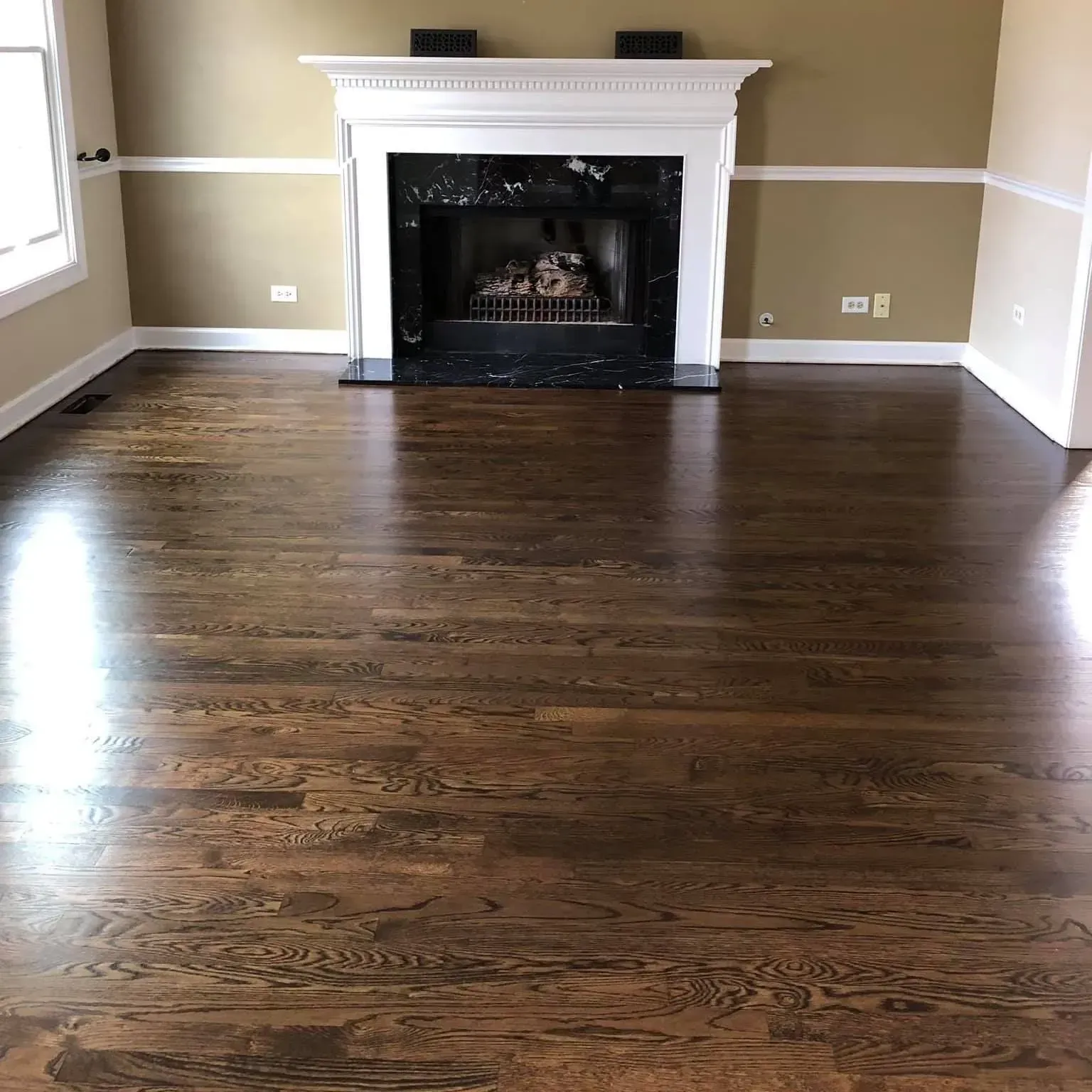 An empty room with dark wood flooring, tan walls, white trim, and a white fireplace mantel with a black marble surround.