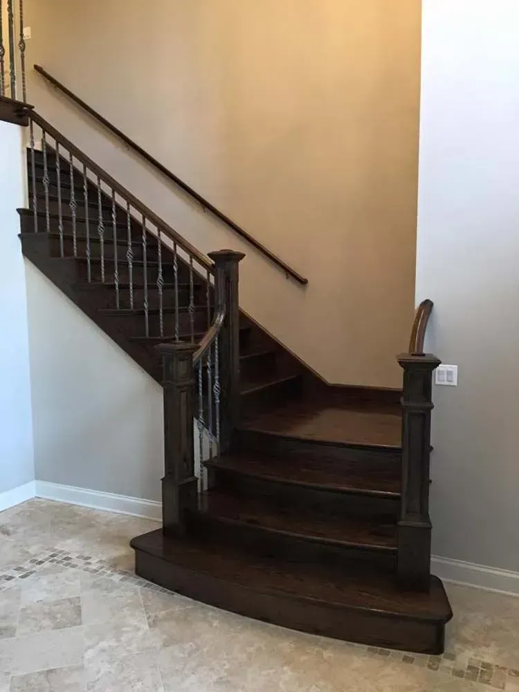 A wooden staircase with metal balusters, a curved bottom step, and dark-stained handrails in a light-walled interior.