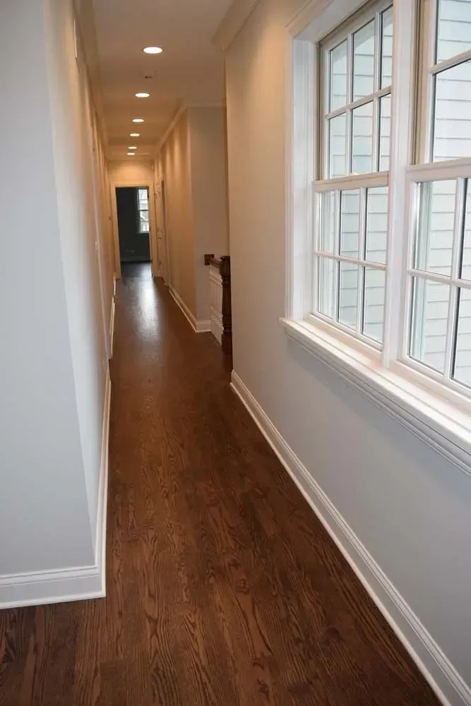 A long, narrow hallway with dark wood floors, light grey walls, recessed ceiling lighting, and a large window.