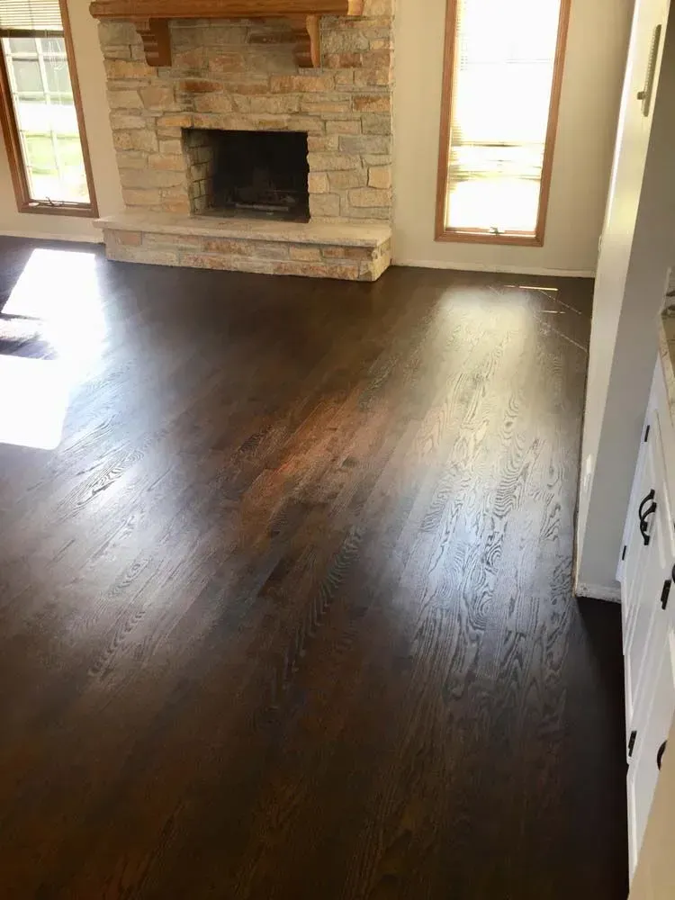 A room with dark-stained hardwood floors, a stone fireplace with a wooden mantel, and two tall windows on either side.