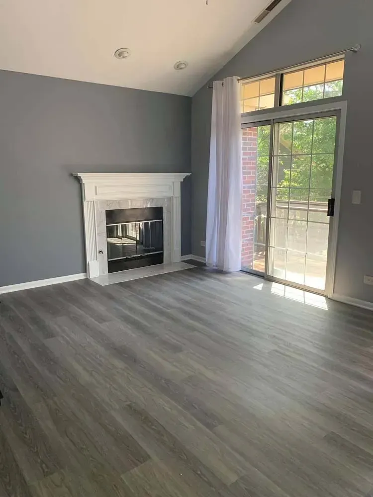 An empty living room with gray walls, wood-look flooring, a white fireplace, and a sliding glass door with a white curtain.