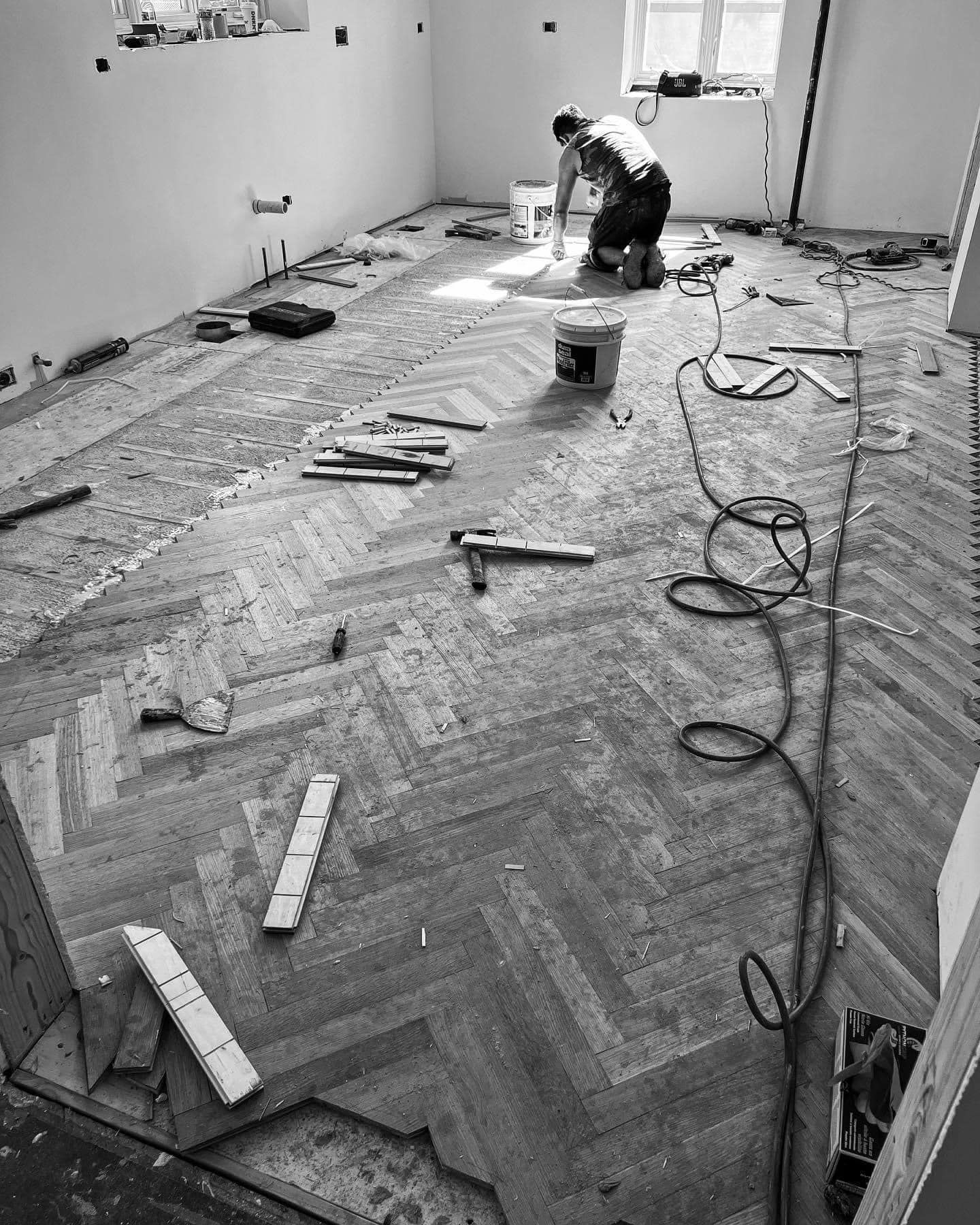 A worker installs a herringbone wood floor in an unfinished room scattered with tools and materials.