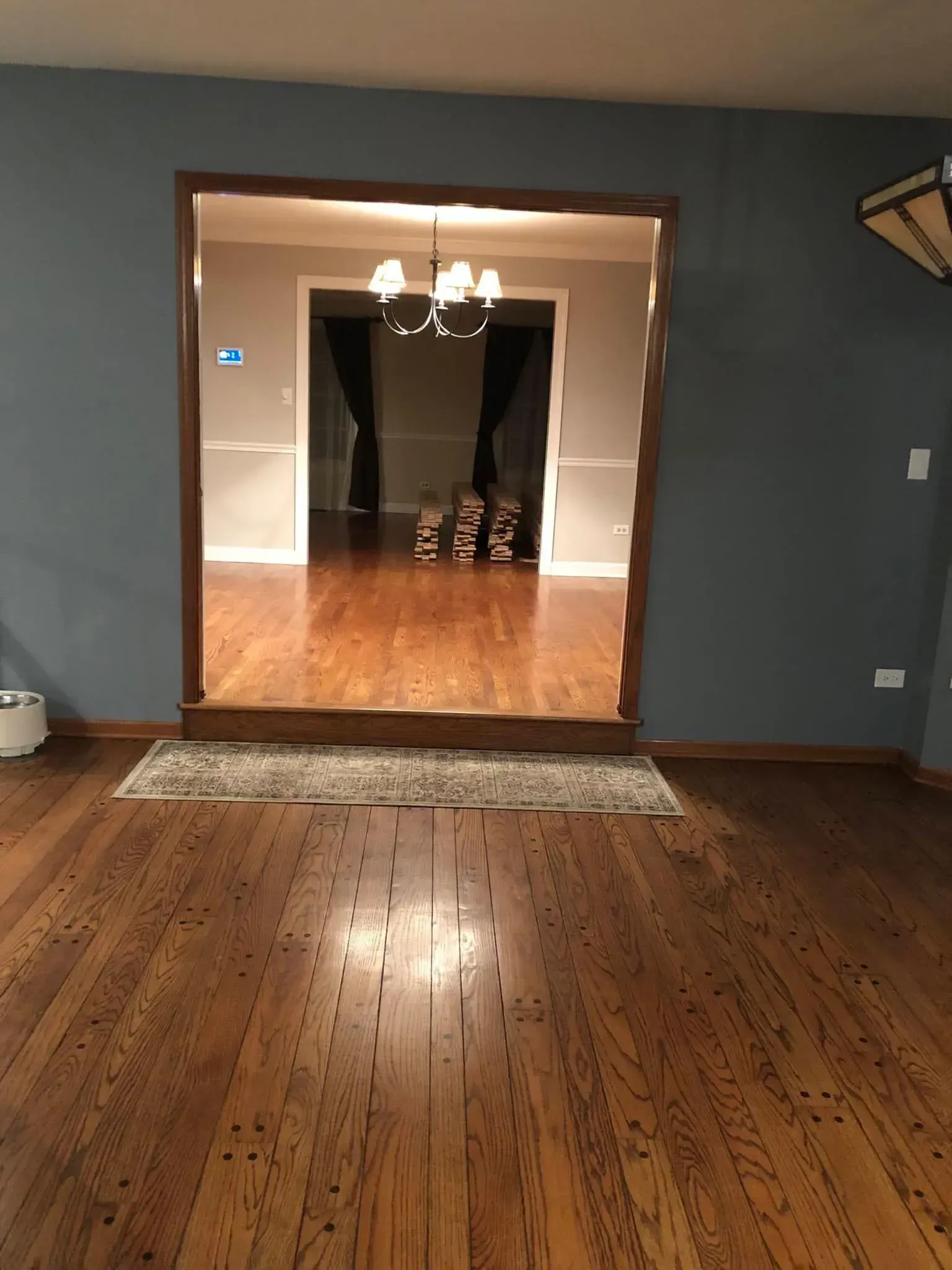 A view through a wooden doorway into an adjacent room with a chandelier, hardwood floors, and dark curtains.