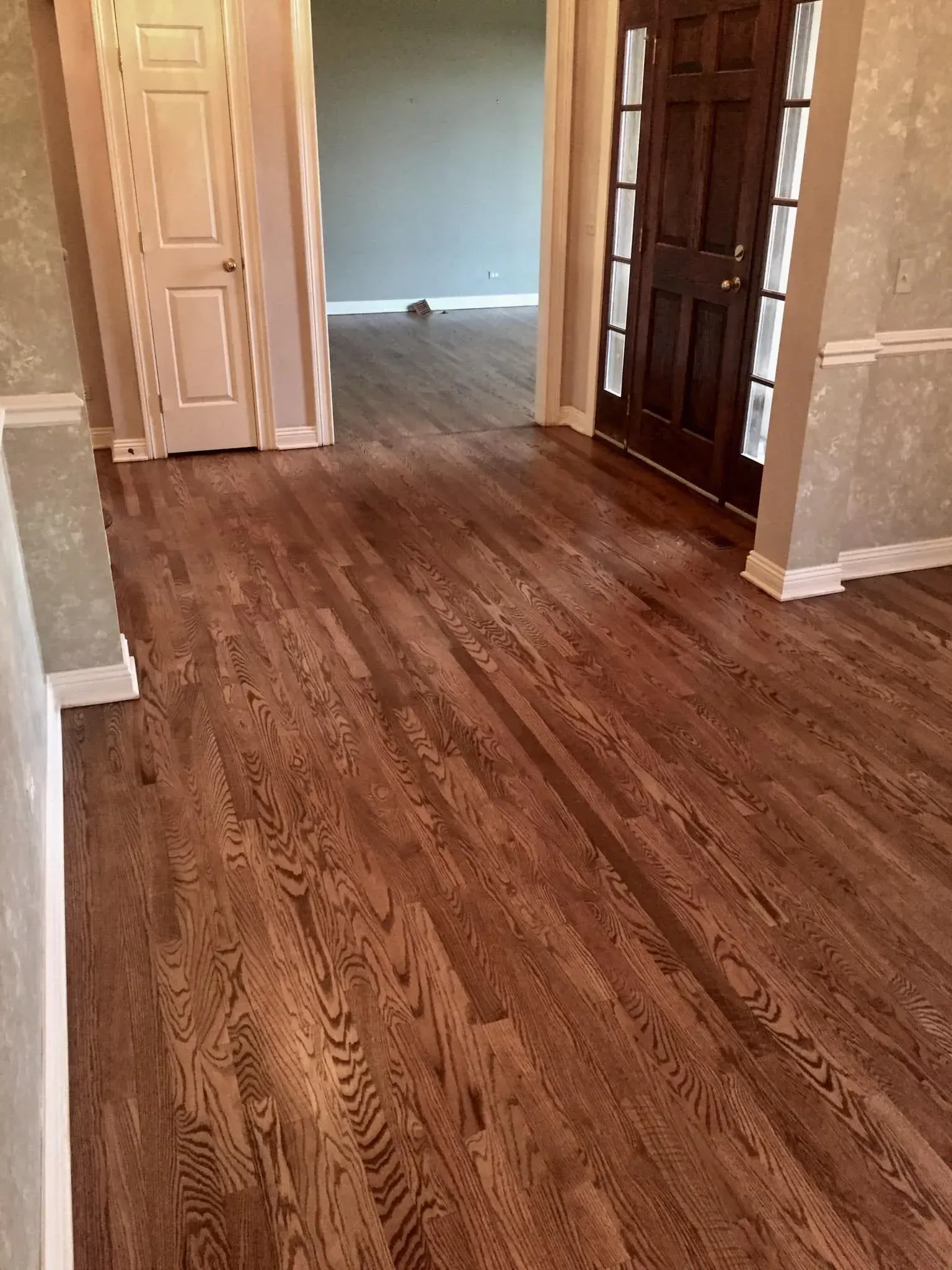 An interior view of a hallway featuring rich brown hardwood floors, a white door on the left, and a dark wooden front door.