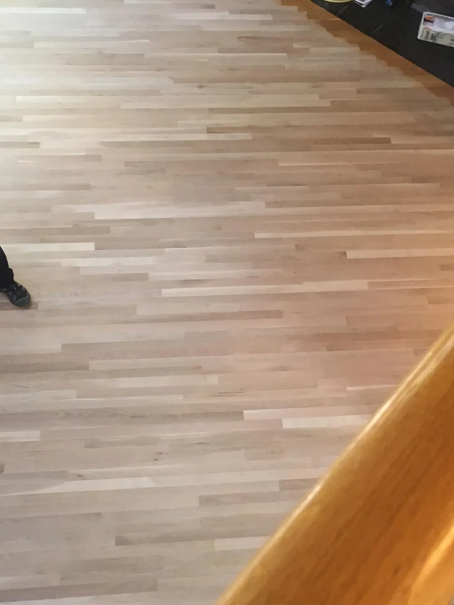 High-angle shot of light-colored hardwood flooring with a person's foot visible on the left and a wooden railing below.