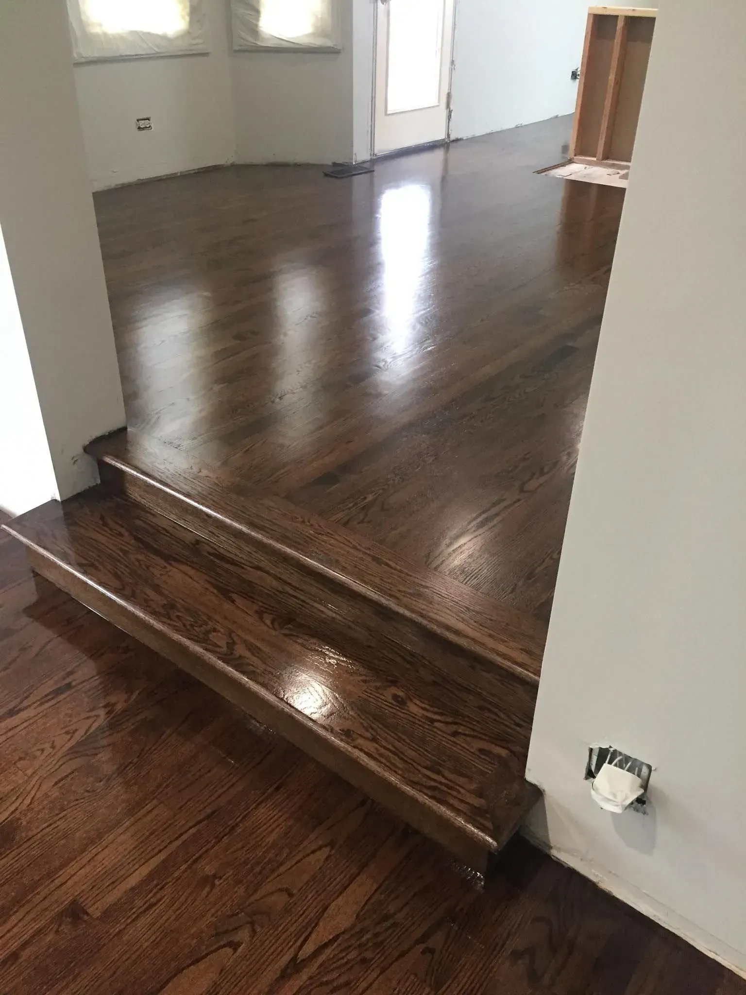 A view from a lower floor looking up two steps at newly finished, dark stained hardwood floors in a sunlit home interior.