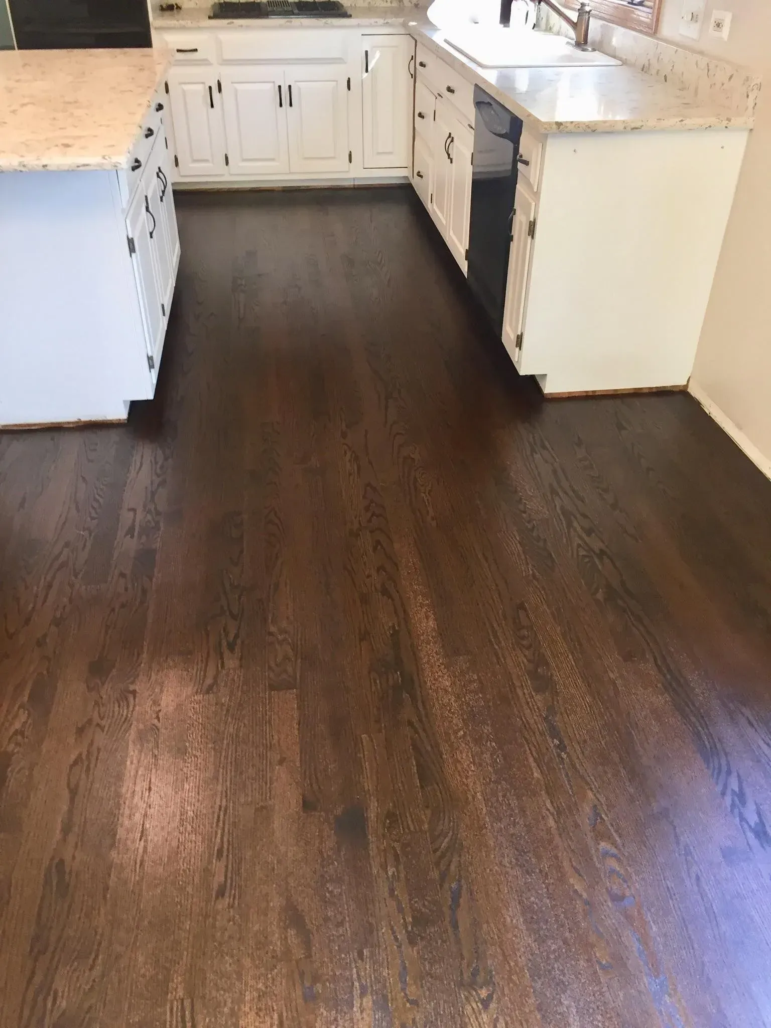 A kitchen floor featuring dark stained hardwood planks, surrounded by white cabinets and a kitchen island with stone tops.