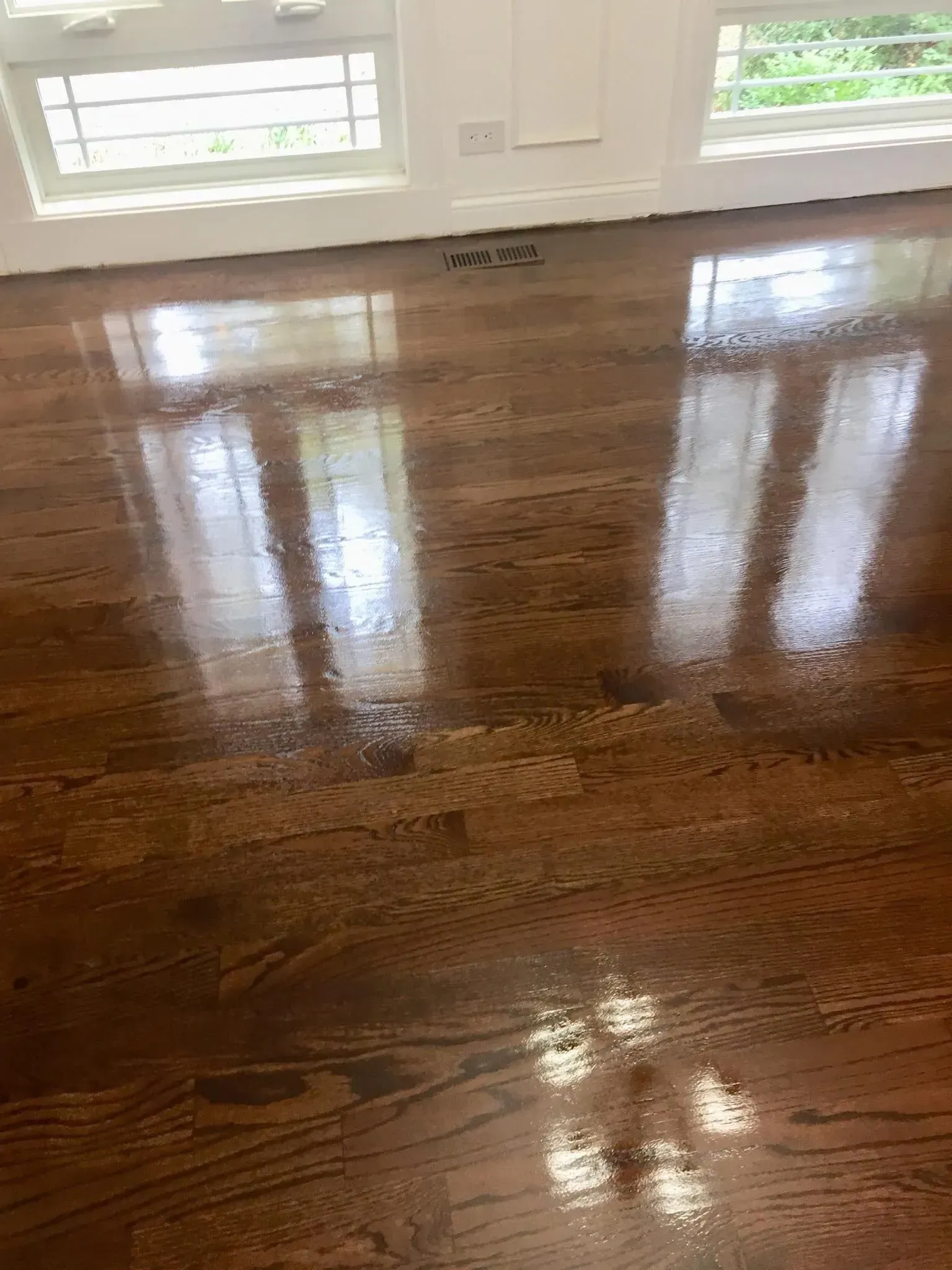 A high-gloss, dark-stained wooden floor reflecting light from two large white windows and a nearby ceiling light.