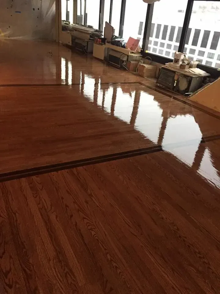 Polished, dark wood flooring reflects large office windows, showcasing a smooth, glossy finish in an empty workspace.