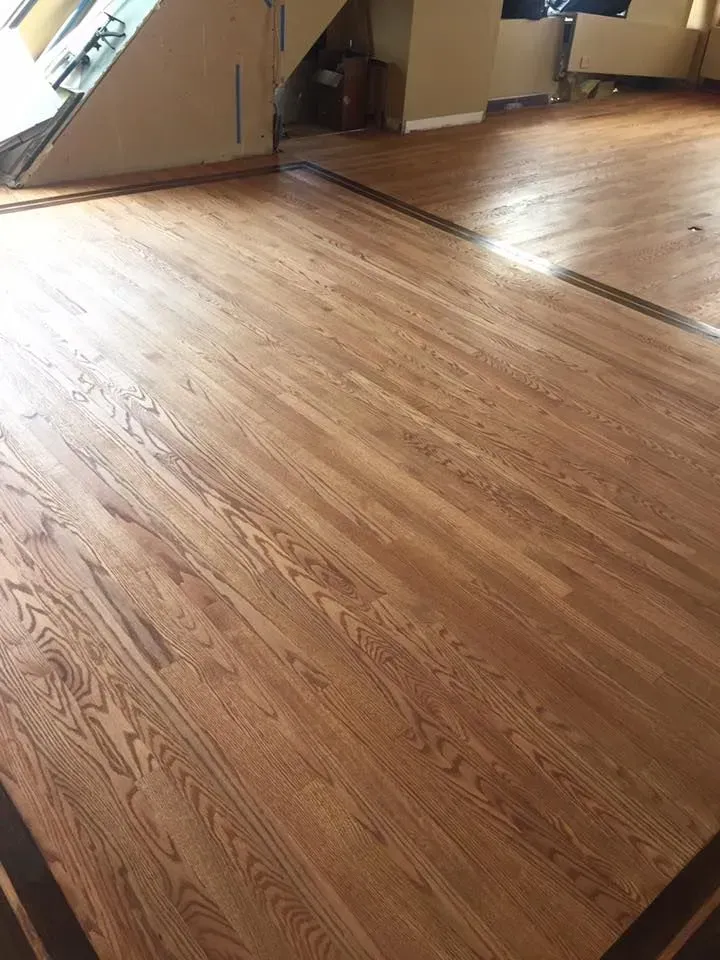 A close-up of light brown hardwood flooring with a dark wood inlay border in an unfinished interior room.