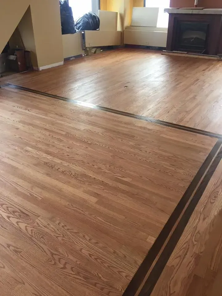 A spacious room with light oak hardwood floors accented by a dark wood inlay border and a fireplace in the background.