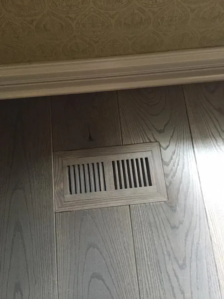 A rectangular floor vent made of wood matches the surrounding light-toned wood plank flooring near a wall baseboard.