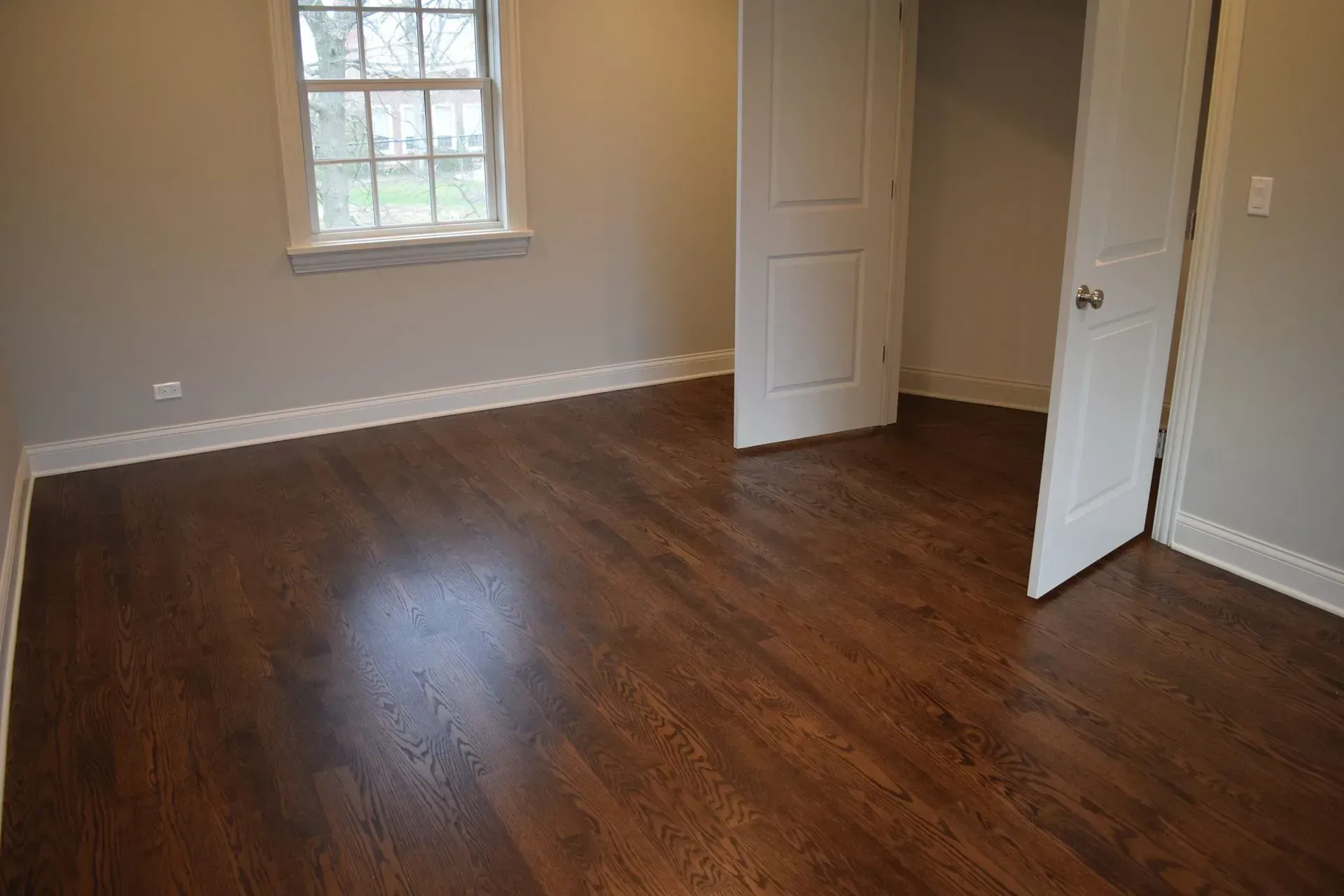 An empty room with dark wood flooring, light grey walls, white trim, a window, and open closet doors.