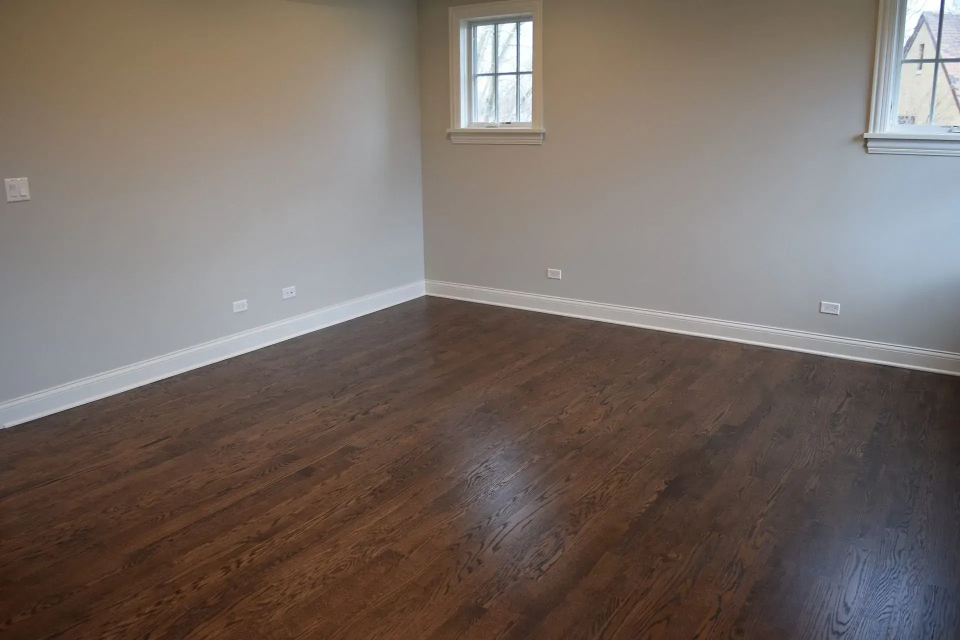 An empty room with light gray walls, white trim, and dark wood floors, featuring two windows on the back wall.
