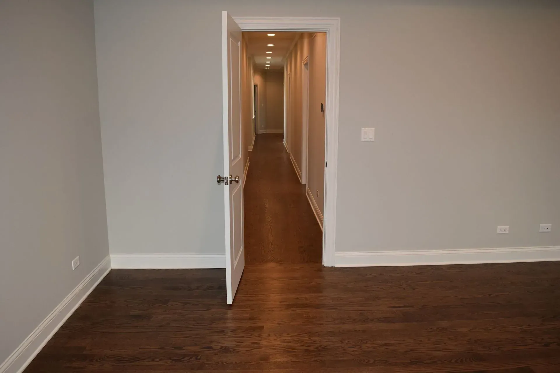An open door in a room with light grey walls and dark hardwood floors leads to a long, dimly lit hallway.