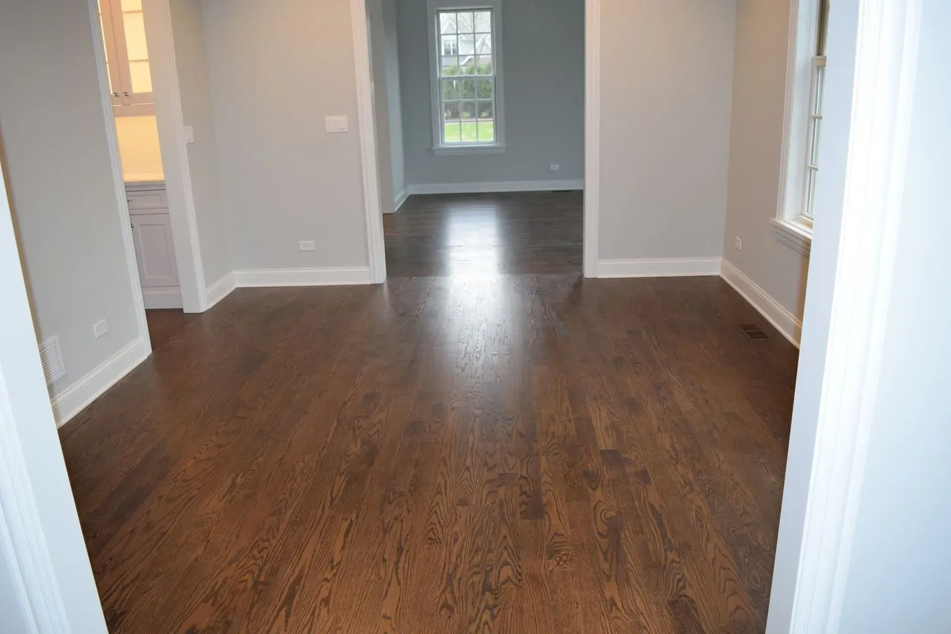 A room with dark wood flooring, light grey walls, white trim, and a doorway leading to another room with a large window.
