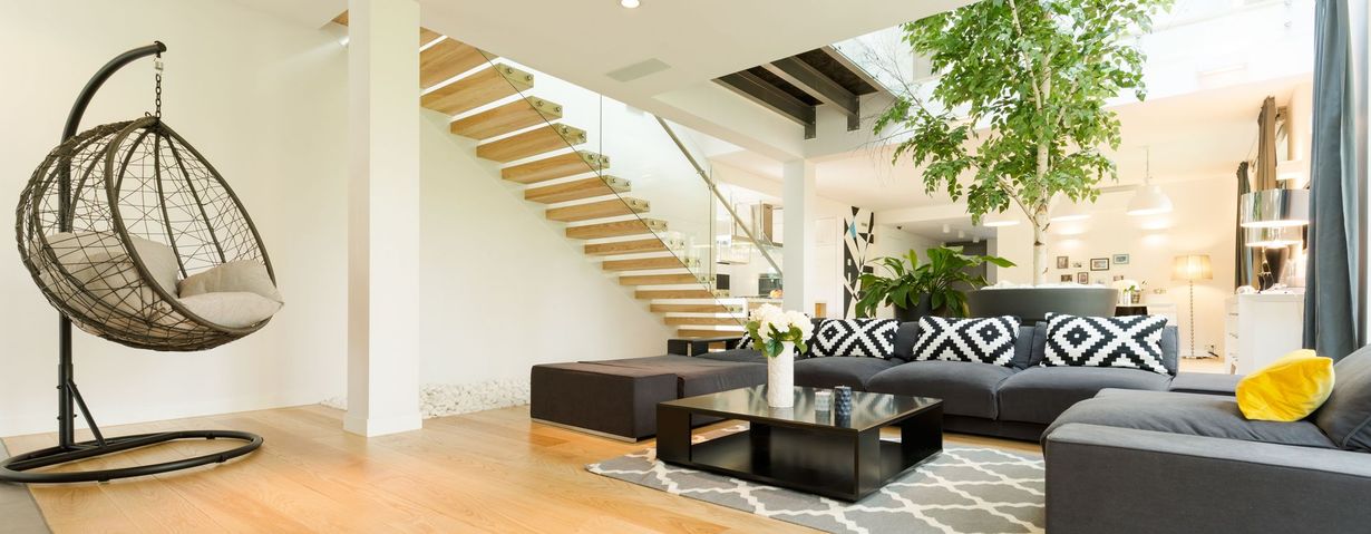A modern, open-plan living room with a black hanging chair, wooden floating staircase, and a large indoor tree.