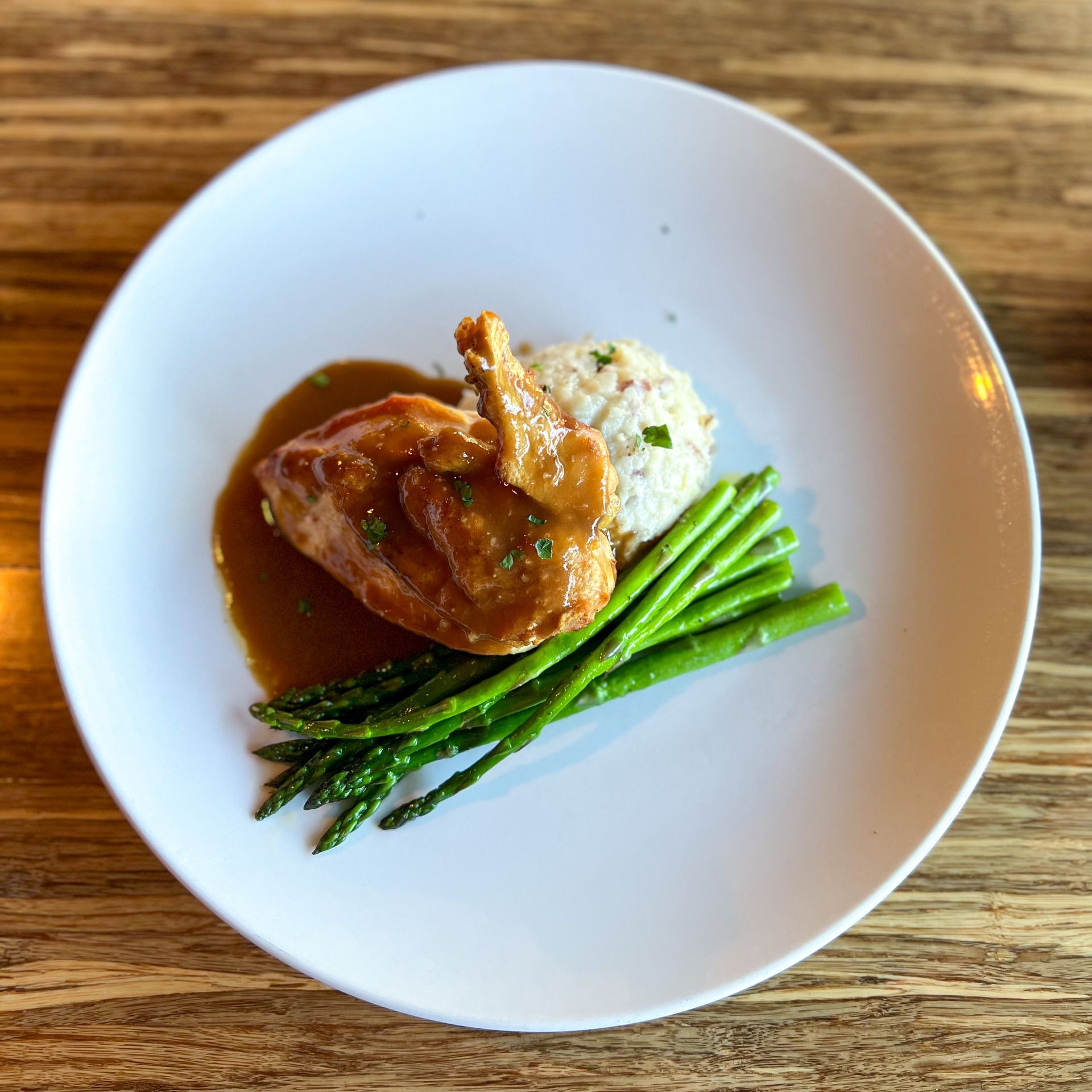 Plated airline bone in chicken breast covered in brown chicken jus. It is served with garlic mashed potatoes and a pile of fresh green grilled asparagus.