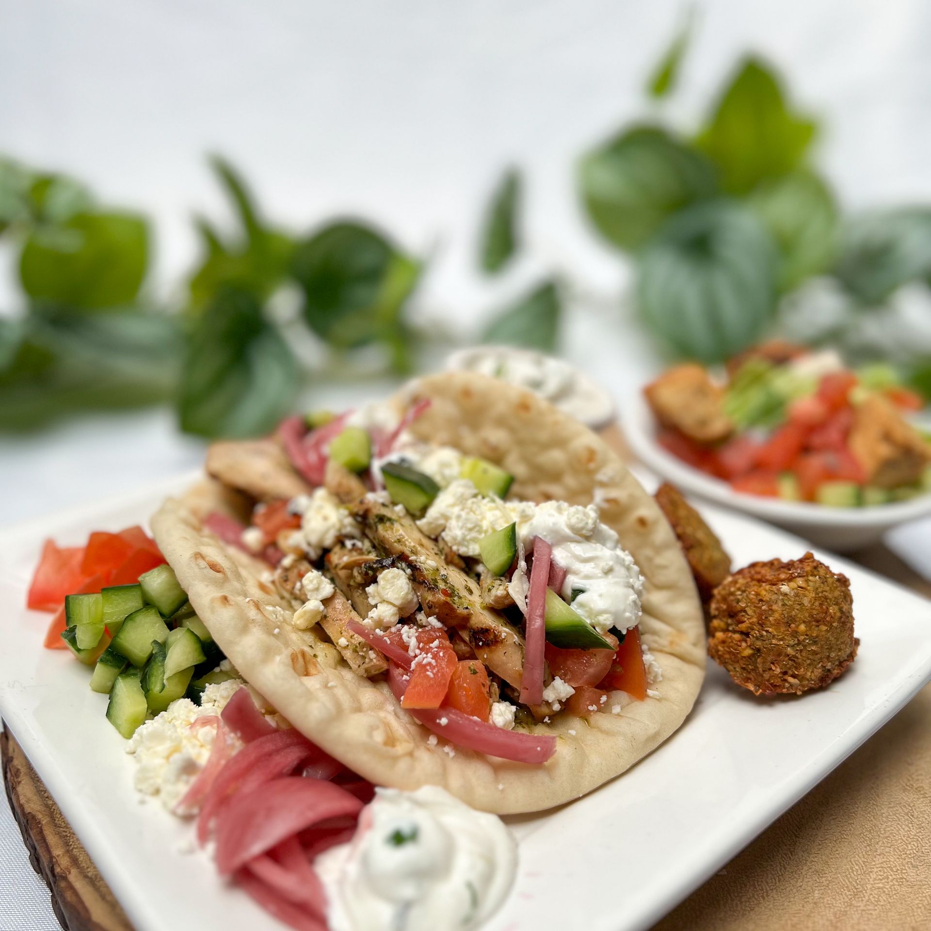 Pita buffet is plated on a white square plate. Pita is stuffed with chicken, fresh tomatoes and cucumber, pickled red onions, and tazikti sauce. There are falafels beside it. 