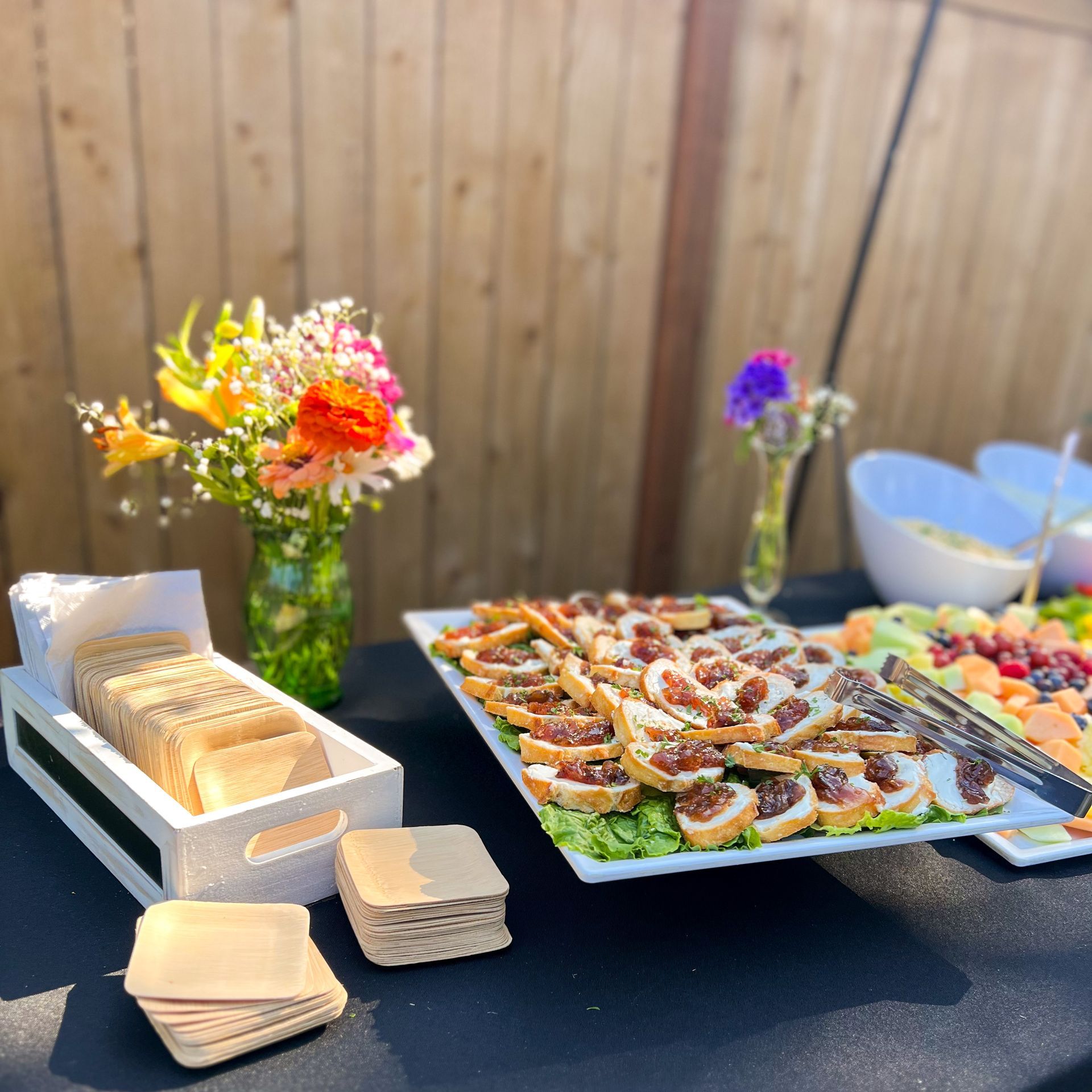 Fig and Goat cheese crostini plated on a platter at a wedding. Black table cloth and brightly colored flower bouquet beside the platter. A stack of compostable bamboo plates is stacked to the left of the platter. 