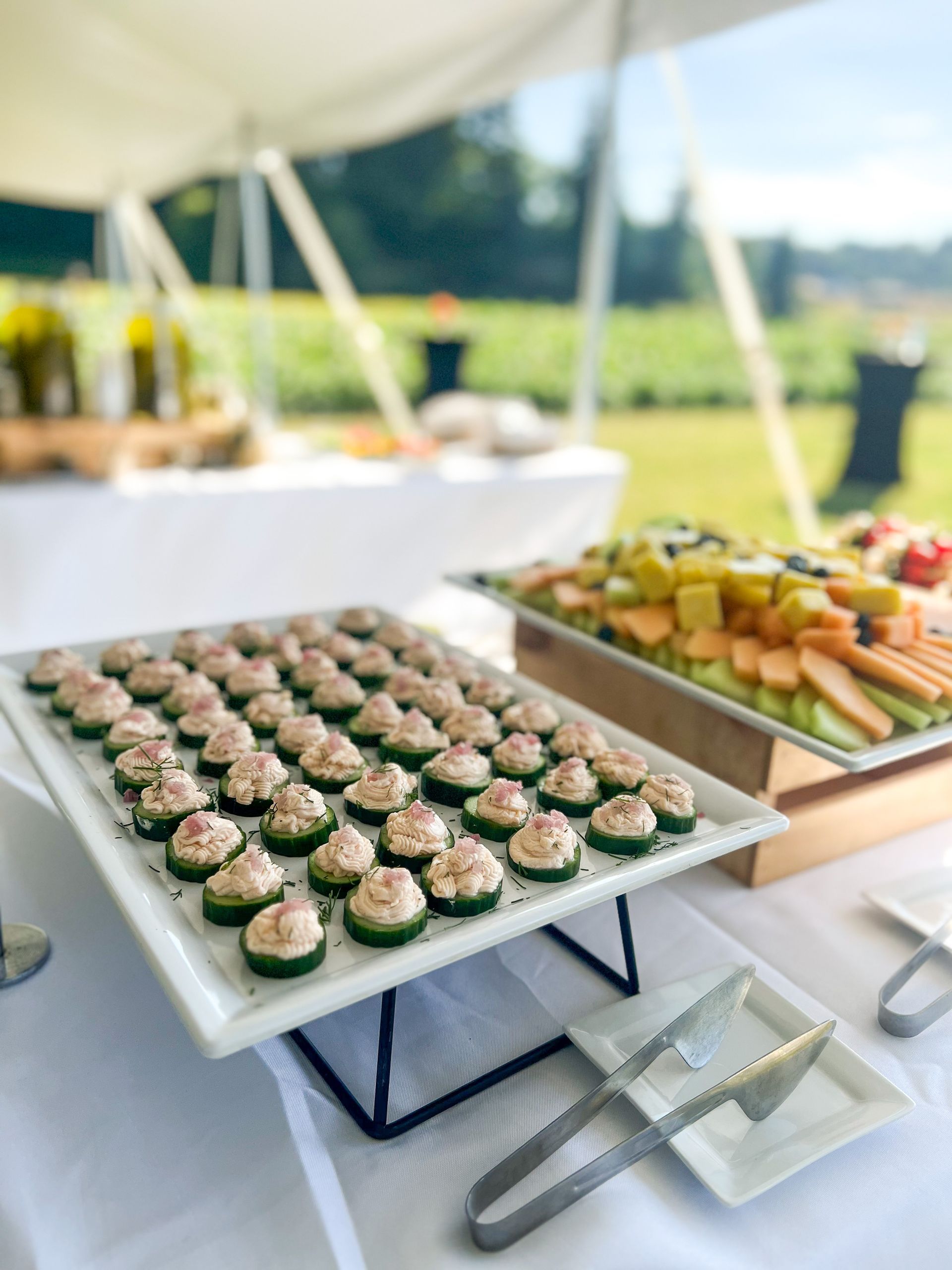 A platter of perfectly lined up cucumber crostinis on a platter. They are on a riser on a buffet with a fruit platter beside them. The table is under a white tent with greenery in the background.
