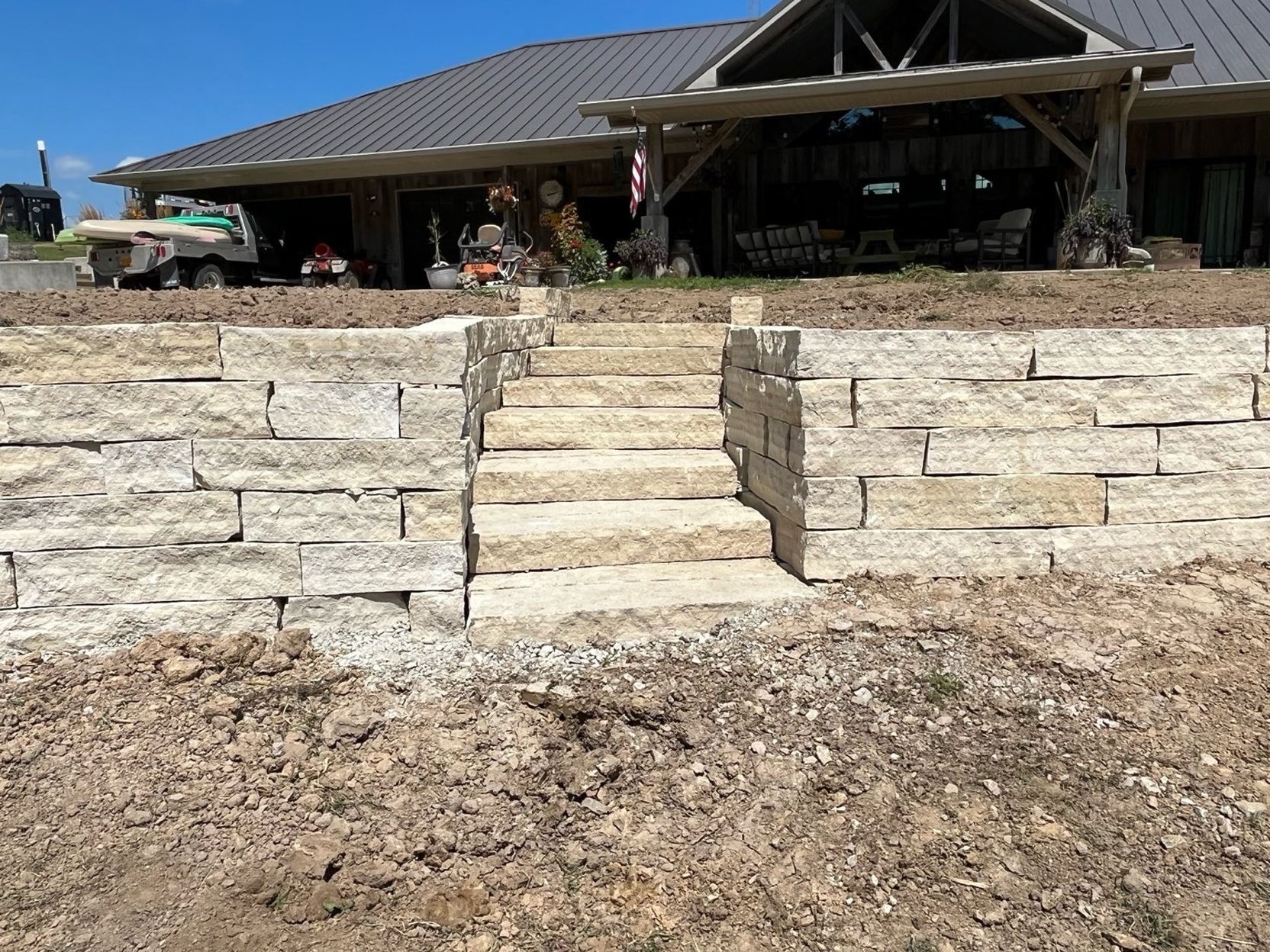 Stone retaining wall with central steps leading up to a house with a covered porch and parked vehicles.