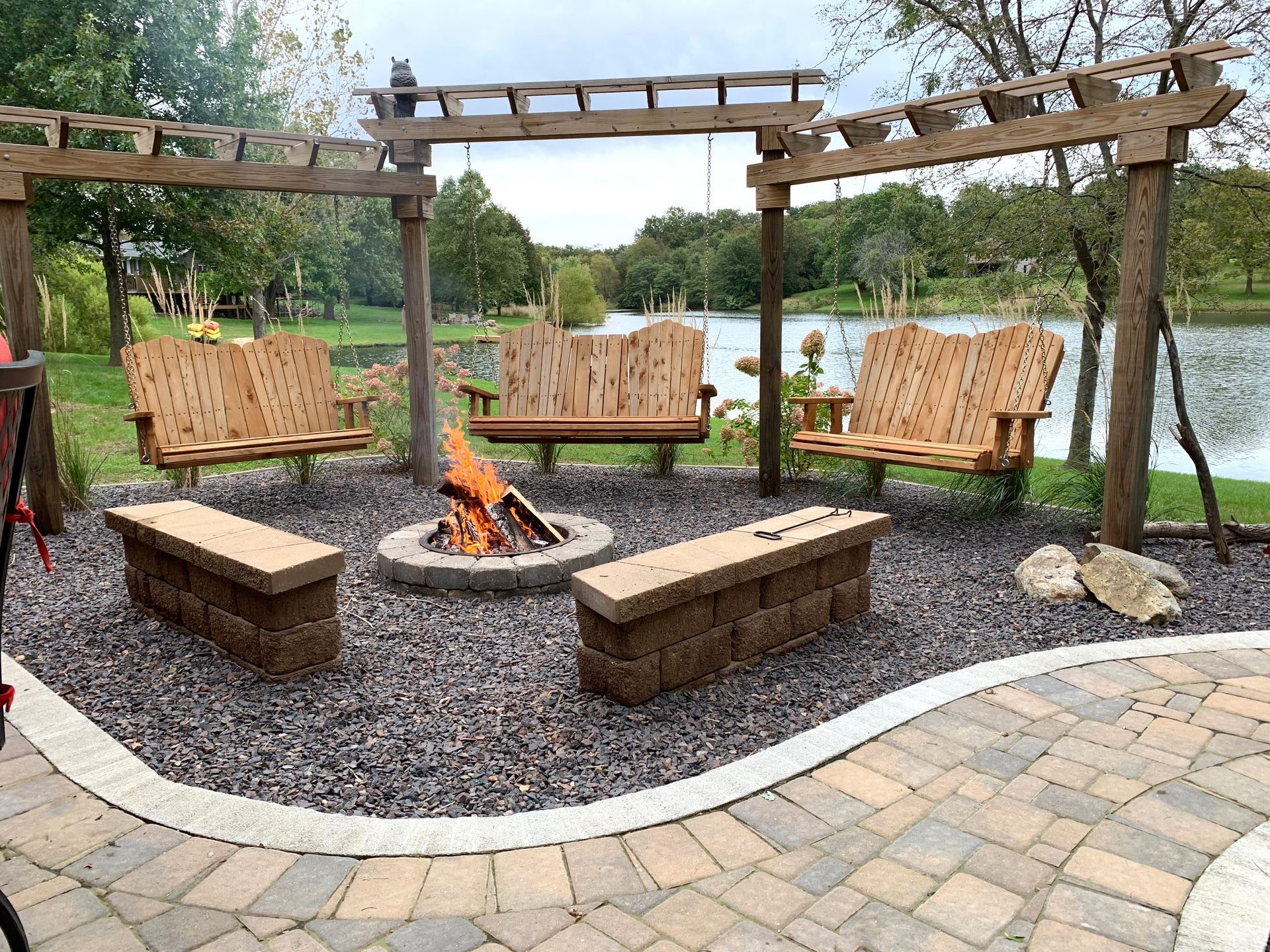 Wooden pergola seating area with benches around a small fire pit on gravel, beside a paved patio and pond.