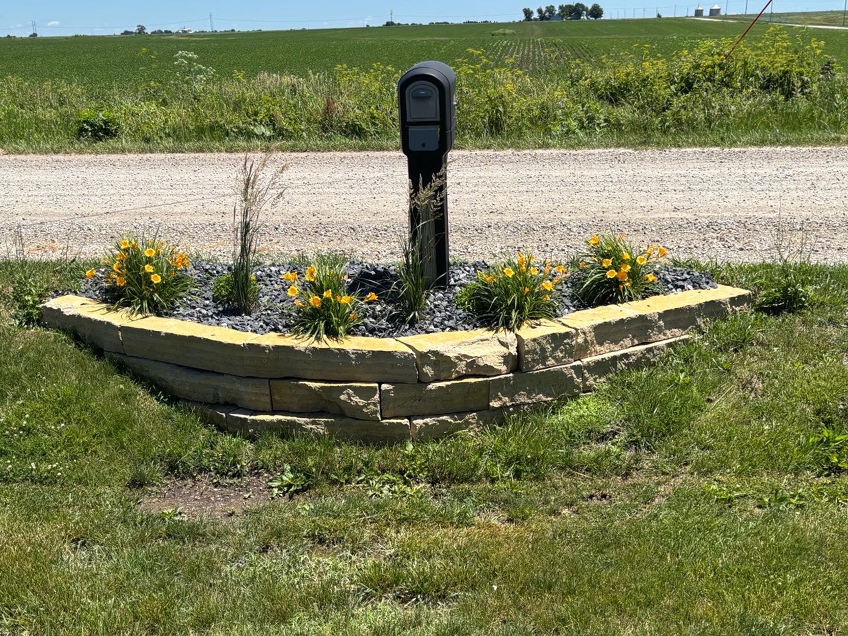 Black post sign in a stone flower bed beside a gravel road and grassy field